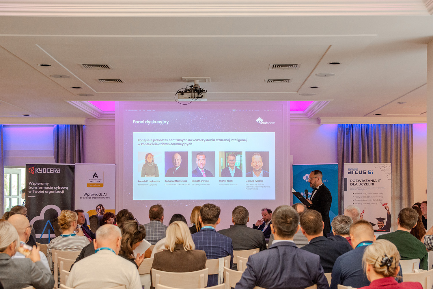 Pełna sala konferencyjna; na ekranie slajd panelu dyskusyjnego z sylwetkami pięciorga prelegentów.